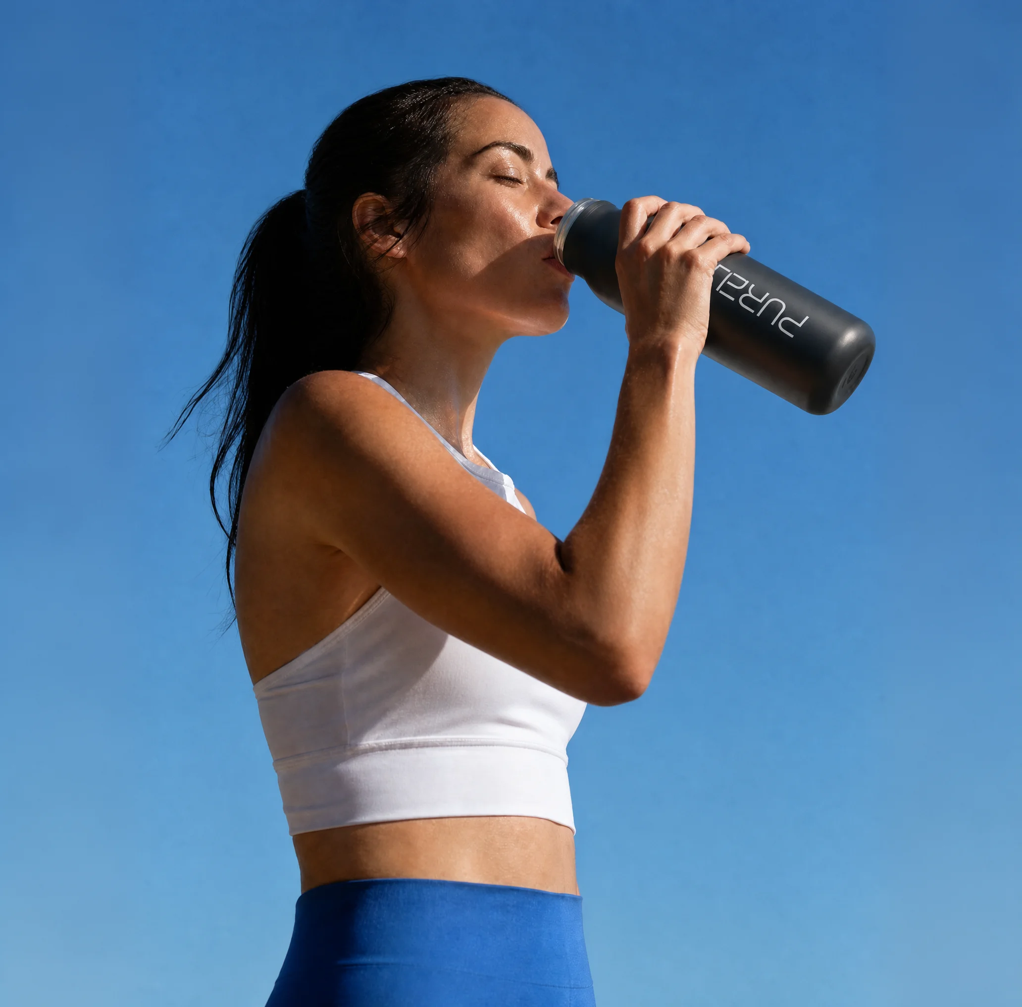 A woman in a white sports bra drinking from a black Purely water bottle against a clear blue sky