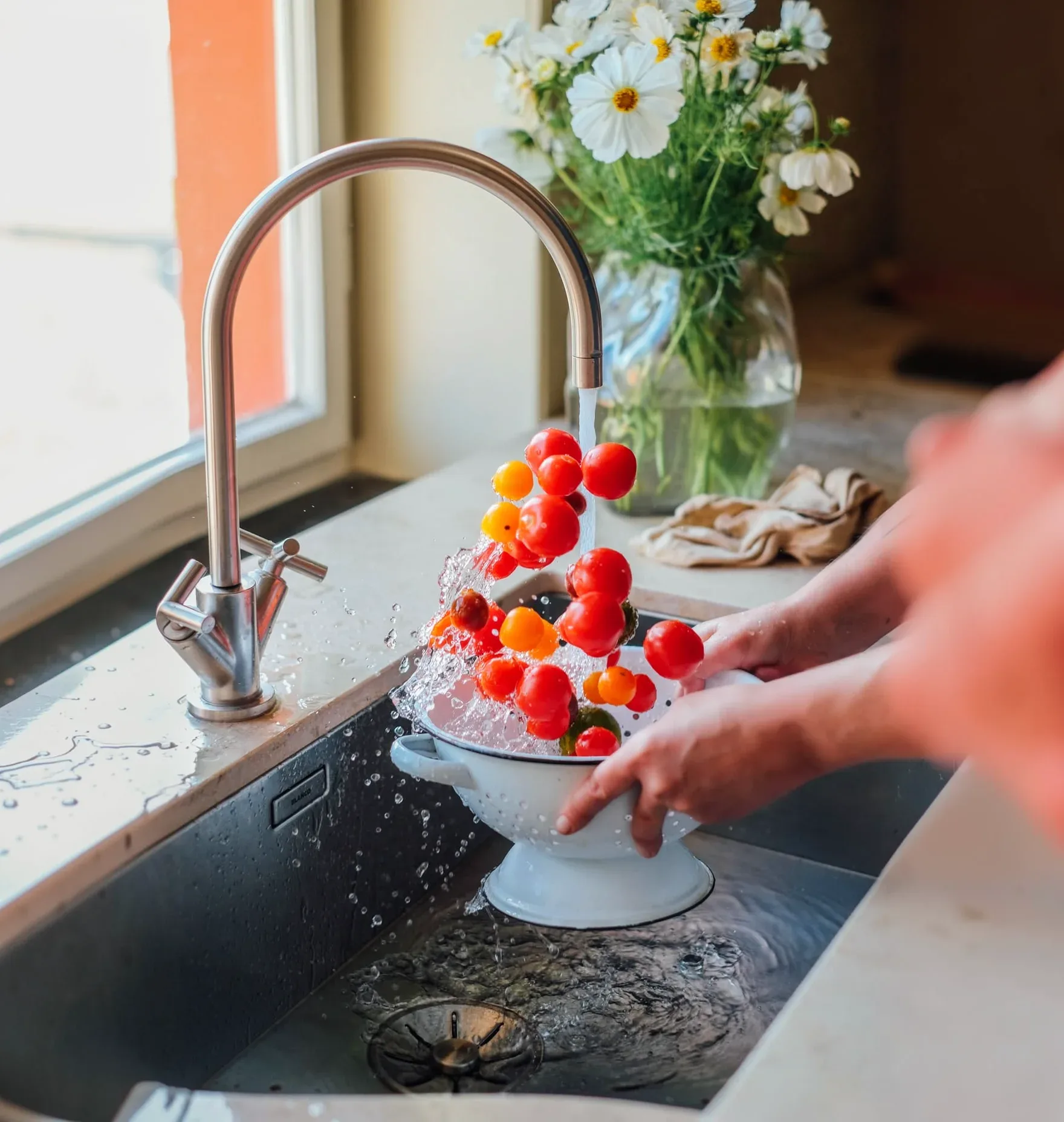 Hands rinsing red and yellow cherry tomatoes in a white colander under a kitchen tap