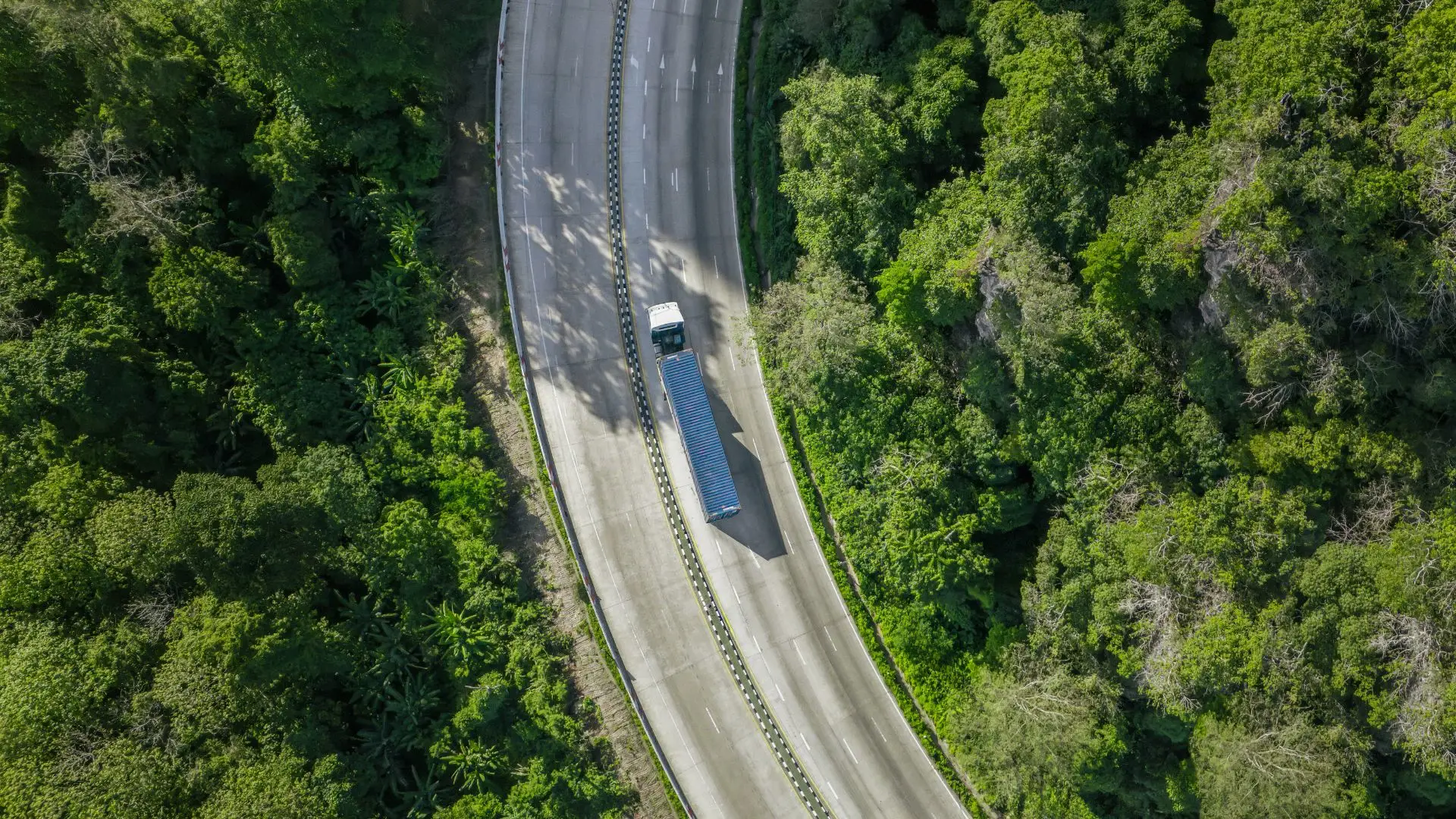 An aerial view of a truck driving along a winding road through dense green forest.